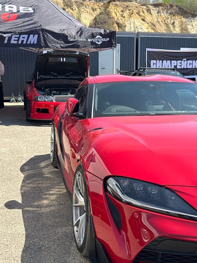 The image shows a close-up view of the front of a red Toyota Supra parked outside a building in Sadovoe, Russia. The car is facing towards the right of the image. Another red car is visible behind it with its hood open. A black tent with white text "TEAM" and "Powered by IMPORTGROUP" is to the left of the cars. A building with a banner containing Cyrillic script is located behind the tent. A cliff face is visible in the background.