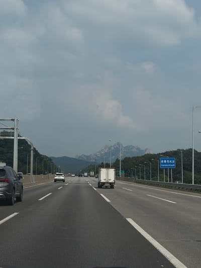 A vertical, eye-level shot captures a multilane highway on a cloudy day, looking towards a mountain range. The highway is paved with asphalt and marked with white lane dividers. Cars and trucks are visible on the road, traveling in the same direction. In the foreground, the left lane has a dark gray SUV. A white box truck is in the middle lane. A blue road sign with Korean and English text is mounted on the right side of the highway, indicating "Gongneungcheon 4 Geore" and "Gongneungcheon4geore". Trees line the sides of the highway, and in the distance, a range of rugged, grey mountains with patches of green vegetation rises against the cloudy sky. The lighting suggests it is daytime.