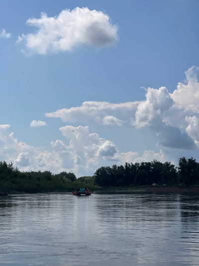 The image depicts a wide, calm river with a tree-lined bank in the background. Several people are visible in a raft on the water, with some holding paddles. The sky is filled with numerous white, fluffy cumulus clouds, and small dark specks, possibly birds, are scattered across the sky. The lighting suggests it is daytime. The scene appears to be a recreational activity on a natural body of water.