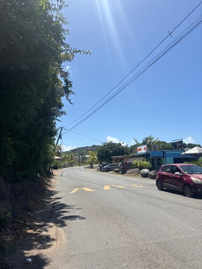 The image captures a sunny street scene in Spanish Town, British Virgin Islands. A paved asphalt road, marked with faded yellow chevrons, curves gently to the left through the center of the frame. To the left, a dense wall of green foliage lines a dirt shoulder where minor debris is visible. A utility pole stands partially obscured by the foliage, with multiple power lines extending across the clear blue sky.
On the right side of the road, a red compact car, likely a Hyundai i10, is parked facing away from the viewer; its front passenger-side tire appears deflated. Behind the vehicle and further down the road, several commercial structures are visible. One building is painted light blue/turquoise and features a partial sign, possibly reading "BAMBU". Adjacent to it, another building presents an extended wooden or thatched canopy. Multiple large, smooth grey boulders are positioned in front of these establishments. Additional vehicles and residential structures are visible in the distance, set against a backdrop of verdant hills under the clear sky. No individuals are present in the scene.