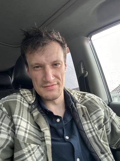 This is a selfie taken by a person inside a vehicle, likely a car, in Makiv, Ukraine. The person, a man with short, dark, slightly damp hair and a light stubble, is looking directly at the camera with a neutral expression. He is wearing a dark blue collared shirt underneath a green and white plaid jacket.
The setting appears to be a cloudy or rainy day, as indicated by the water droplets visible on the car's windows and the overcast sky through the sunroof. The interior of the car is visible, including part of the dashboard and the roof lining. The image is taken from a slightly low angle, with the man's head and upper body filling most of the frame. There is no visible text in the image.