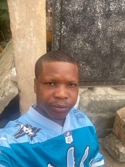 In Ojo, Nigeria, a medium shot captures an adult male with dark skin and a short haircut, directly facing the camera with a neutral expression. He wears a light blue and turquoise mesh athletic shirt featuring a white stylized bird logo on the left shoulder and a white NFL shield logo with "MYA" text on the chest. Partial text "GOD'S KINGDO" is visible on the upper right chest.
The background consists of two main vertical sections. To the left, a light brown, weathered concrete or plaster pillar stands alongside grey rubble and a glimpse of green foliage. To the right, a dark, possibly metal, surface is heavily marked with irregular white streaks and speckles. Below this dark surface, more grey concrete or stone is visible, and in the bottom right corner, a partially open cardboard box with indistinct contents and partial text ("BEAR...", "N.A.C...") rests on the ground. The scene is illuminated by natural light.