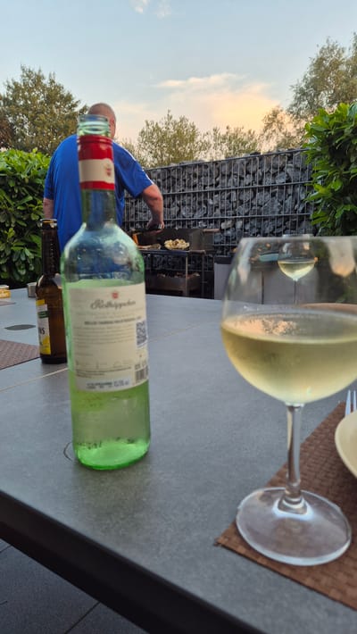 A man is grilling outdoors on a patio in Dissen am Teutoburger Wald, Germany. He is wearing a blue shirt and glasses. There are two bottles of drinks on the table, one green glass bottle and one brown glass bottle. There is also a glass of white wine. In the background, there is a stone wall and trees. The sky is clear with a few clouds.