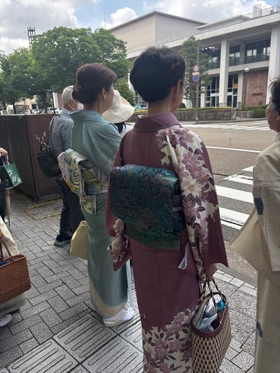 The image captures a street scene in Kanazawa, Japan, showing several individuals standing on a sidewalk, primarily viewed from behind, under a cloudy sky.
In the mid-ground, three women are dressed in traditional Japanese kimonos. The central figure wears a light teal-colored kimono with a subtly patterned fabric. Her white obi (sash) is decorated with a vibrant pattern of green, yellow, and blue stylized leaves. Her dark hair is styled in a neat, low bun. To her right, a woman is seen in a deep reddish-purple kimono. This kimono features prominent white panels on the sleeves and lower back, adorned with patterns of purple and white flowers and dark green leaves. Her obi is a dark, iridescent teal with a rich, textured appearance. She holds a rectangular, dark brown woven basket bag with fine horizontal gold stripes in her right hand. Both women wear white tabi socks and white zori sandals. To the far right, partially visible, is a third woman in a light cream-colored kimono with a delicate hexagonal geometric pattern and a small, rectangular blue patch on the sleeve.
Behind the woman in the teal kimono, an older man with grey hair is visible. He wears a short-sleeved grey collared shirt, dark pants, and carries a black shoulder bag. Another individual, largely obscured by the central figure, wears a wide-brimmed white sun hat. On the far left, a hand is partially visible holding a green paper bag with a white graphic.
The group stands on a grey paved sidewalk composed of rectangular blocks, interspersed with tactile paving blocks featuring parallel ridges. Across a street, marked by a white pedestrian crossing (zebra stripes), a large, modern, light-colored building with multiple stories is visible. This building features a prominent colonnade and large windows. Green trees are interspersed throughout the background, partially obscuring other structures. A red circular traffic sign indicating "30" is mounted on a pole near the street, accompanied by a smaller blue rectangular sign. A black street lamp with a white globe is also visible across the street.