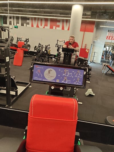 The image displays an indoor gymnasium in Nahariya, Israel, captured through a large wall-mounted mirror. In the central reflection, a Caucasian male with short light-colored hair and visible tattoos on his right arm is seated on a black gym machine. He wears a red t-shirt and white over-ear headphones, holding a dark-colored smartphone in both hands and looking at its screen. The gym machine he uses features a digital display showing exercise diagrams and text, including "rollcallader 1/84". To his right, a large white cylindrical pillar extends from floor to ceiling. The background reveals additional gym equipment, including weight machines with red upholstered pads, exercise benches, and weight racks, all situated on a dark rubberized floor. A white wall behind the reflected man displays large red lettering partially visible as "WE LOVE LIF". Overhead linear lighting fixtures are reflected in the ceiling. In the immediate foreground, the perspective originates from a red upholstered gym equipment pad with white stitching. A digital display screen, part of this foreground equipment, is visible, showing workout instructions with multiple body movement diagrams and the text "rollcallader 1/84". A crumpled light green towel rests on the dark floor to the right of the red pad. Further reflections in a distant mirror on the far right show two additional individuals in dark attire.