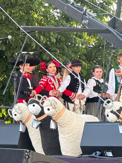 The image depicts a group of young performers on a stage, dressed in traditional folk costumes. In the foreground, three large, knitted sheep figures with bells around their necks are positioned on top of large black speakers. Behind the sheep, from left to right, are several individuals. The first two are children in what appears to be mariachi attire, featuring wide-brimmed hats, jackets with ornate detailing, and sashes. The child on the right is a girl wearing a vibrant red dress with intricate embroidery and red flowers in her hair. Next to her is a boy in a black vest over a white shirt, with decorative stitching on his trousers. To their right, a girl wears a dark vest over a white blouse and a long grey skirt, and another individual to her right is dressed in a white shirt with a green vest. The background consists of lush green foliage, suggesting an outdoor setting. The stage appears to be set up for a performance, with microphones and cables visible. The presence of a stage, speakers, and performers suggests a cultural event or festival. Considering the context of traditional folk costumes and potential performance, this scene is likely taking place during a cultural celebration in Nowy Sącz, Poland, possibly at an outdoor venue or park hosting such events.