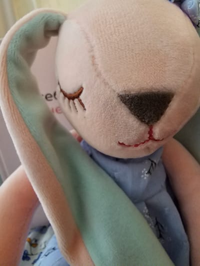 A close-up, angled shot focuses on a plush toy, possibly a bunny, with a light beige body and mint green inner ears and trim. The toy is adorned with embroidered features, including a dark grey triangular eye outlined with red stitching, and a stitched mouth resembling an "X" shape. The toy is wearing a light blue garment with a subtle pattern of white flowers and small blue dots. The background is soft and blurred, with hints of peach and pink tones suggesting an indoor setting. The lighting appears to be natural, with a gentle glow. No specific time of day, weather, or location cues are discernible, but the plush material and the clothing suggest a domestic environment. The overall impression is one of a child's toy, possibly being held or displayed. The visible text on a tag, partially obscured, reads "er" and "ie". The image is devoid of any human subjects.