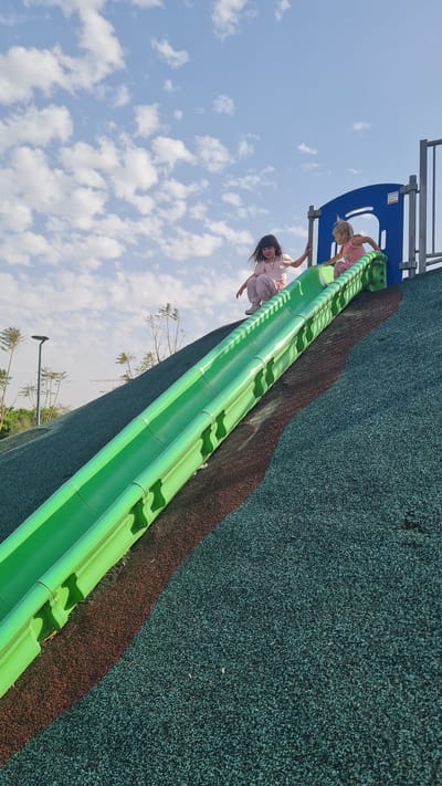 Two girls are on a green slide at a playground in Kiryat Gat, Israel. The younger girl, with blonde hair and wearing pink, is at the top of the slide, while the older girl, with dark hair and also wearing pink, is sliding down. The slide is adjacent to a blue and gray playground structure, and the ground is covered in dark green and brown safety surfacing. The sky is visible in the background with scattered white clouds. Trees and streetlights are visible at the bottom of the slide.