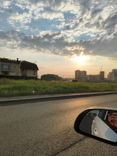 This media file captures a view from inside a moving vehicle on a road, likely in Металлплощадка, Russia, as indicated by the location context. The scene is set during sunset, with the golden light of the setting sun casting a warm glow on the clouds and the asphalt.

In the foreground, the road surface is visible, reflecting the sunlight. To the left of the road, a grassy field stretches towards a building that appears to be a two-story house or a small apartment building, with a dark-tiled roof and light-colored walls. Beyond this building and across the field, a line of trees forms a backdrop. Further in the distance, a construction site is visible with several tall buildings under construction, including one with a prominent crane. The sky is filled with dramatic clouds, some white and fluffy, others darker and more textured, with the sun peeking through them.

In the side mirror of the vehicle, the reflection shows a white car and a hand holding a phone, suggesting that the image was taken by someone using their phone to capture the scene. The overall mood is peaceful and serene, indicative of a late afternoon or early evening drive. There are no visible text elements in the image.