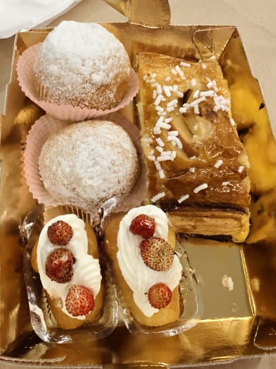 A golden rectangular cardboard box contains a variety of pastries. The box is partially filled, with a white paper liner visible in the upper left corner. The pastries are arranged in a somewhat disorganized manner.
In the upper left quadrant of the box are two round pastries dusted with powdered sugar. They are placed inside pink paper cupcake liners.
To the right of the powdered sugar pastries is a rectangular pastry with a flaky crust. It is glazed and topped with white sprinkles, and appears to contain sliced fruit, possibly apples, underneath the glaze.
In the bottom half of the box are two small, elongated pastries topped with white cream and small, whole strawberries. These pastries are resting in clear plastic containers.
The background is a light brown surface. The image appears to have been taken from a slightly elevated angle, looking down into the box of pastries.
The image does not contain any specific visual information that would allow for the precise identification of its location within Pozzuoli, Italy.