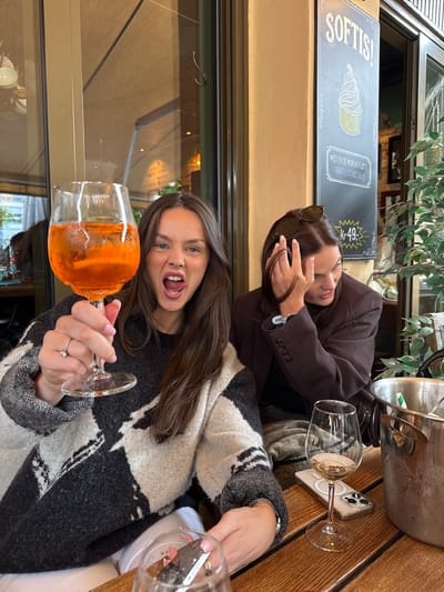Two young women are at an outdoor table. One is holding a glass of an orange liquid, possibly Aperol spritz. She has brown hair, blue eyes, and is sticking her tongue out. She is wearing a patterned black and white sweater. The other woman is wearing a brown jacket and sunglasses in her hair. The setting appears to be a restaurant in Drammen, Norway, as indicated by a sign in Norwegian. A metal ice bucket and an iPhone are on the table. One glass is empty and the other has a little white liquid left in it.