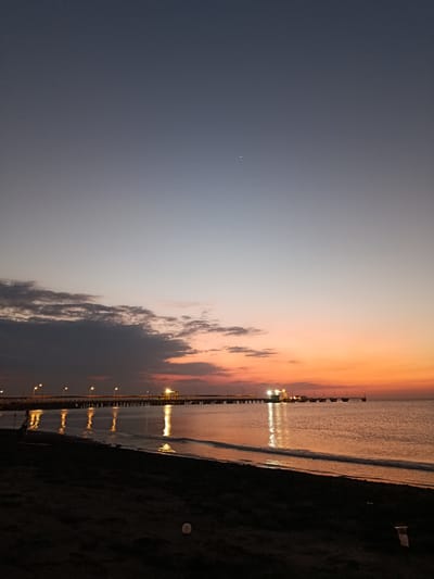This media file depicts a tranquil twilight scene at the coast in Jangkar, Indonesia. The foreground shows a dark, sandy beach with some scattered debris, including a plastic bottle and a piece of wood. To the left of the frame, a solitary figure can be seen standing on the beach, adding a sense of scale and human presence to the vastness of the scene.
The main subject of the image is the expansive ocean meeting the sky. A long pier stretches across the water, supported by numerous pilings, and is illuminated by a line of streetlights. At the end of the pier, there appears to be some industrial or port infrastructure, with brighter lights suggesting activity. The lights from the pier reflect shimmering golden paths on the calm surface of the sea.
The sky transitions from a deep indigo at the top to warm hues of orange and pink near the horizon, indicating the time of day is either sunrise or sunset. A few scattered, dark clouds are present in the mid-sky, contrasting with the otherwise clear gradient of colors. A faint crescent moon is visible in the upper left portion of the sky.
The overall mood of the image is peaceful and serene, evoking a sense of calm as day transitions into night. The stillness of the water and the gentle glow of the lights create a contemplative atmosphere. There is no visible text in the image.