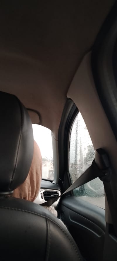 Inside a car, a view from the back seat showcases a rainy window. The seatbelt is fastened. The headrest of the front seat is black leather. The window is speckled with raindrops, blurring the outside view. Part of the front passenger is visible, wearing a soft, tan-colored sweater. A ventilation vent is visible on the dashboard.