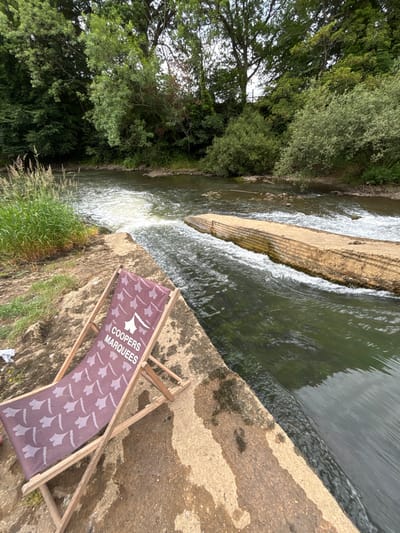 A sideways view depicts a river with a small waterfall cascading over a tiered concrete structure. Lush green trees line the riverbank. A weathered concrete surface stretches into the foreground. On this surface rests a purple and white beach chair with the text "COOPERS MARQUEES" printed on it. The chair is slightly reclined. The scene suggests a tranquil, riverside setting with a touch of commercial branding.