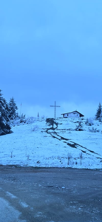 The image captures a serene winter landscape, rotated 90 degrees clockwise from its natural orientation. When viewed correctly, a clear, pale blue sky dominates the upper portion of the frame. Below, a snow-covered hillside slopes upwards from left to right, with a dirt or gravel road running along the bottom.

In the mid-ground, nestled on the snowy hill, stands a small, white-walled building with a dark roof, resembling a chapel or a small house. To its left, a prominent, large metal cross rises from the snow, its structure clearly visible against the blue sky. Further down the hill and to the left, another, smaller cross or marker can be faintly seen. Distinct dark tracks are etched into the snow on the hillside, suggesting recent passage.

The landscape is sparsely populated with trees, many of which are evergreens heavily dusted with snow, particularly noticeable on the left side and foreground. A flag pole, adorned with a red and white flag (possibly Polish or Indonesian), is visible in the lower-left foreground, partially obscured by snow-laden branches.

The scene depicts a cold, overcast day, with diffused light illuminating the snow, giving the entire image a cool, blueish tint. There are no people visible, and no specific activities are taking place, lending a quiet, peaceful, and perhaps solemn atmosphere to the setting. No visible text is present in the image. The location is Stoikite, Bulgaria, which aligns with the mountainous, snowy terrain depicted.