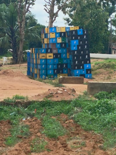 Outside of Zonkwa, Nigeria, a disorderly stack of blue, yellow, and black beer bottle crates dominates the foreground. The crates are irregularly arranged, rising above the sandy, dirt landscape. A stand of trees, including palm varieties, flanks the stack on the left. Behind the crates, the scene transitions to a green area with a building visible in the distance on the right. The lower portion of the image depicts an uncultivated area with bare earth and scattered green vegetation.