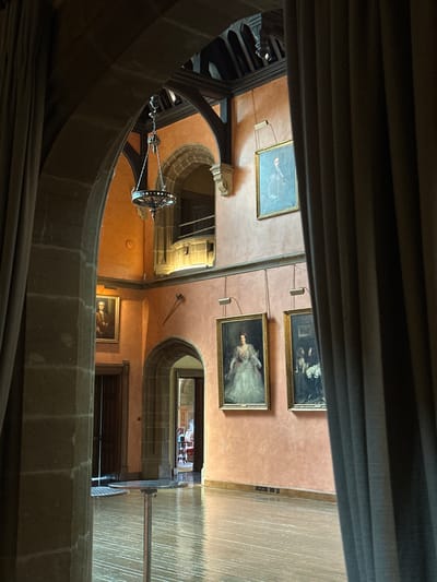This is an interior shot of a grand hall with a warm, peach-colored wall. The hall is dimly lit, with light streaming in from an unseen source, creating a reflective sheen on the polished wooden floor. A large, dark archway frames the view into the hall, with heavy, brown curtains partially obscuring the foreground.
On the peach wall, there are several framed oil paintings. In the upper left, a painting depicts a man in historical attire. To its right, another painting shows a woman in a formal gown. Below these, a third painting features a woman in a light-colored, flowing dress, seated on a chair. To the right of this, a fourth painting shows a man and a dog in a dimly lit setting. Hanging above the paintings are small, adjustable spotlights.
A medieval-style chandelier hangs from the vaulted, dark wooden ceiling. The hall features stone architectural elements, including arches and columns. Two doors are visible, one dark wood door on the left and another, more recessed, doorway to the right, leading further into the building.
The scene is a static, interior view of a formal hall, possibly within a historic building. The location is not specified in the image itself, but the description does not contain any information to suggest it is located in Chichester, United Kingdom.