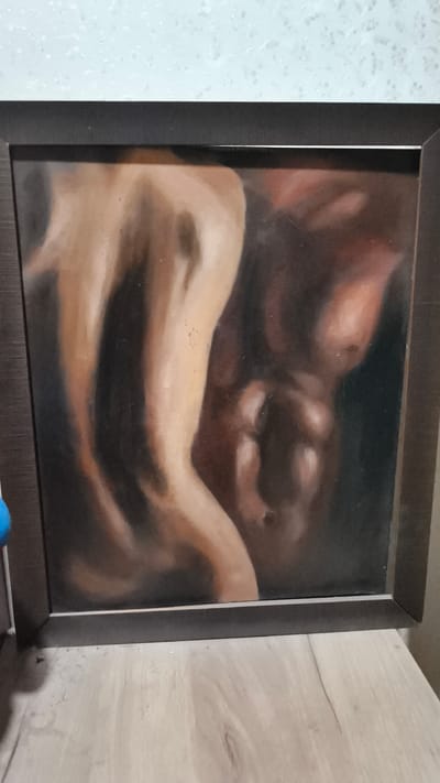 The image shows a framed painting of a human torso, likely male, with detailed musculature rendered in oil paint. The painting is positioned on a wooden surface, possibly a shelf or table, in front of a textured, light-colored wall. The framing is dark and appears to be wood. The scene is indoors. There is no discernible location in the city of Мирзо Турсунзода, Tajikistan.