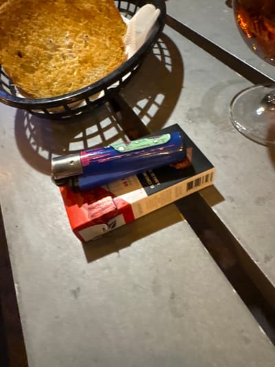 A top-down view shows a table setting. A bread basket with toast sits to the left. Next to it is a blue lighter with cartoon-like images on it, resting against a red-and-white box. A partially visible wine glass and other container sit at the top of the image. A bar with a shadow stretches horizontally along the top right. The table's surface is mottled gray.