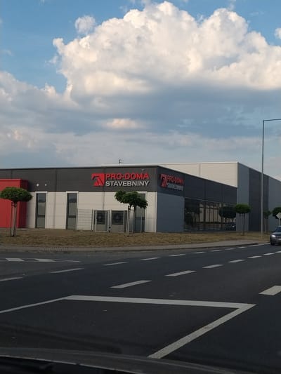 This is a vertically oriented, eye-level shot of a modern building with the text "PRO-DOMA STAVEBNINY" prominently displayed in red and white lettering. The building appears to be a commercial establishment, possibly a hardware or construction supply store, given the "STAVEBNINY" part of the name, which translates to "building materials" in Czech. The architecture is sleek, featuring dark grey and white panels, large glass windows, and what appear to be air conditioning units on the exterior.
In front of the building, there is a strip of dry grass and two small, ornamental trees in planters. To the right of the building, a paved road is visible with white lane markings, suggesting it's a public thoroughfare. A dark-colored car is driving away from the camera on this road.
The sky above is a mix of blue and white clouds, indicating daytime. The lighting suggests it might be late afternoon or early evening, as the light is somewhat diffused. The scene is set in Horažďovice, Czechia. The overall impression is one of a functional, modern commercial area on a day with mixed cloud cover. There are no people visible in the image.