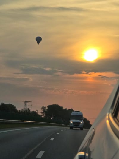 The image captures a sunset scene on a highway in Lazdėnai, Lithuania. A hot air balloon is visible in the sky, floating above a brightly lit sun. In the foreground, a road with white lane markings curves to the right. A white van is driving in the same direction as the viewer, in front of another vehicle, which is partially visible on the right side of the frame. Trees and a power line tower can be seen on the left side of the highway. The sky is filled with clouds, tinged with orange and yellow hues from the setting sun.