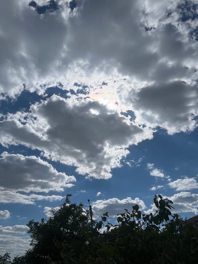 This is a vertical, eye-level, medium shot photograph of a cloudy sky with the tops of trees in the foreground. The sky is a vibrant blue with numerous white and gray clouds scattered throughout. Some of the clouds are dark and appear to be heavy with rain, while others are lighter and fluffier. The sun is partially obscured by clouds, but its rays are peeking through, creating a bright, ethereal glow. There is a faint rainbow visible in the upper left portion of the sky. In the foreground, the dark silhouettes of trees are visible against the bright sky. The leaves of the trees are dense and appear to be in full foliage. A single street lamp is visible among the trees. The scene is set in Kryzhanivka, Ukraine.