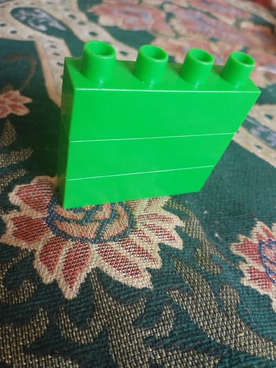 A close-up, eye-level shot shows a bright green, four-stud Lego brick positioned diagonally on a textured, multi-colored rug. The rug has a dark green background with intricate floral patterns in shades of red, cream, and blue. The Lego brick is oriented so that its top surface, featuring four cylindrical studs, is partially visible. The brick's smooth, rectangular sides are also prominent. The lighting is natural, casting subtle shadows. No people are present in the image. The scene is an indoor domestic setting, likely a home in Mady, Kyrgyzstan, given the provided context.