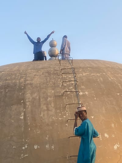 The image displays three males on and around a weathered, light-beige concrete dome structure in Barako, Nigeria, set against a clear, pale blue sky. The dome's surface exhibits numerous cracks and areas of plaster detachment. A vertical series of dark metal ladder rungs are affixed to the dome. In the lower right foreground, a young male, wearing a teal tunic with white trim and a patterned kufi cap, stands on a rung. He looks towards the viewer with a slight smile, his right hand near his temple. At the dome's apex, a second young male, clad in a blue shirt and dark trousers, kneels with both arms extended upwards in a V-shape, fingers splayed. To his right, a third male, in a light-colored tunic, stands with his back mostly to the viewer, positioned near a decorative finial. The finial consists of three stacked, golden-toned spherical elements culminating in a spire.