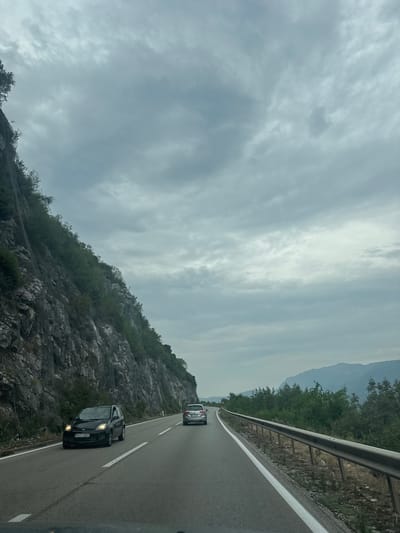 The image shows a road curving through a mountainous landscape with a steep, rocky cliff face on the left side. The cliff is covered with some vegetation and a mesh barrier is visible on its surface, likely for protection against rockfalls. A dark-colored car with its headlights on is driving in the left lane of the road. Further ahead, a silver car is in the right lane. The road is bordered by a metal guardrail on the right, separating it from a dense growth of green trees and shrubs. In the background, hazy mountains are visible under a cloudy sky. The overall impression is of a scenic drive on a cloudy day. The scene is likely located near Kujava, Montenegro, given the rugged terrain and vegetation common to the region.