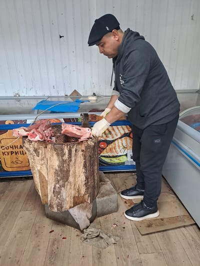 A man in Obukhovo, Russia, wearing a dark grey hoodie, black pants with a Fila logo, black shoes with white soles, and a black cap, is butchering meat on a large wooden block. He is wearing white gloves and using a cleaver. Pieces of meat are visible on the block and around its base. The background includes a cooler or display case, a wall with white paneling, and a floor with wood-patterned laminate. A sign in Cyrillic script is visible at the base of the block.