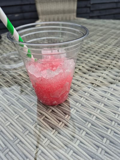 A clear plastic cup filled with pink slushy sits on a woven outdoor table. A green and white striped straw extends from the cup. In the background, a dark wooden fence and a blurred fabric chair are visible. The scene is set outdoors, likely on a patio or deck. There is no specific indication of the location being Carrick-On-Shannon, Ireland.