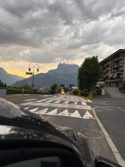 This is a photograph of a road intersection in Saint-Gervais-les-Bains, France, with a mountainous landscape in the background. The image is taken from inside a vehicle, looking forward. The sky is overcast with dramatic clouds, and the sun is setting or rising, casting a warm glow on the distant mountains and sky.
In the foreground, the hood and windshield of a dark-colored car are visible, with raindrops on the glass. The road surface is wet, indicating recent rain. A pedestrian crossing with white triangular markings is prominent in the middle of the frame.
To the left of the road, a street lamp with two lights stands next to a blue sign that reads "GENDARMERIE" with an arrow pointing left. There is also a red yield sign. Further back, a town with buildings and trees is nestled at the base of the mountains. A large, leafy tree is situated on the right side of the road, near a multi-story building with balconies.
A motorcycle with a glowing headlight is visible on the road, approaching the pedestrian crossing. A silver van and a dark-colored car are parked near the building on the right. The overall scene depicts a transition in weather, with the dramatic sky and wet roads contrasting with the serene mountain backdrop.