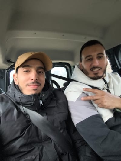 The image depicts two adult males seated inside a vehicle. While the contextual information indicates the city of Sabaa Aiyoun, Morocco, the specific location within the city is not visually discernible from the image itself, which shows an indoor vehicle environment.

The male positioned on the left is wearing a tan corduroy baseball cap and a dark, possibly black, puffer jacket. A black seatbelt is visible diagonally across his chest. He has a light complexion, dark hair, a short mustache, and a short beard, and is looking directly at the camera with a neutral expression.

The male positioned on the right is wearing a multi-colored hooded jacket, segmented into white on the top, light grey in the middle, and dark grey on the lower portion of the arm and torso. He also has a light complexion, dark hair, a mustache, and a short beard. He is looking directly at the camera and has his right hand raised, forming a V-sign (peace sign) with his index and middle fingers extended.

The vehicle interior features a light grey ceiling and dark-colored window frames. The external view through the vehicle windows is indistinct.