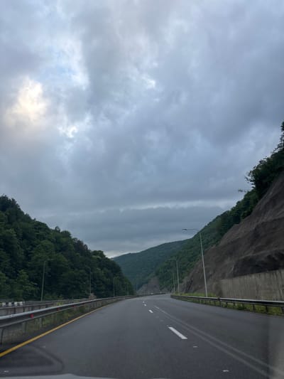 The image depicts a view from a vehicle traveling on a multi-lane asphalt highway, likely in the city of Tsakva, Georgia, given the lush green mountainous terrain. The highway is wet, indicating recent rain. Guardrails line the sides of the road, with yellow and red reflectors visible on the left. The road curves gently ahead, disappearing into a valley. Steep, tree-covered hillsides flank the highway on both sides. On the right, a large, excavated rock face is visible, reinforced with concrete structures and metal netting. Streetlights are positioned along the highway. The sky is overcast with heavy, grey clouds, with a patch of lighter sky with some sunlight peeking through on the upper left. No people are visible in the frame. The scene captures a travel moment on a scenic, mountainous highway under cloudy weather.