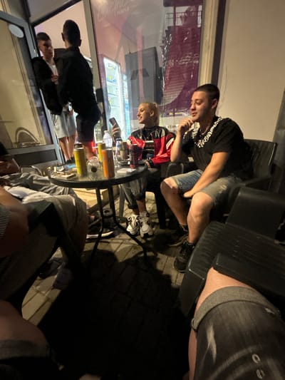 In Krivodol, Bulgaria, at night, several individuals are gathered around a small, round glass-top table outside what appears to be a commercial establishment with a glass-fronted display. Two men are standing near the entrance, one in a black jacket and white shorts, the other in a black jacket and dark shorts. A woman with blonde hair tied back is seated at the table, wearing a red and black jacket and holding a cell phone. Next to her, a man with short hair and a chain necklace is seated, wearing a black t-shirt and denim shorts. Other individuals are partially visible, seated or standing, with various drinks and cans on the table. The ground is paved with light-colored stones, and shadows are cast by the artificial lighting. The establishment's interior features illuminated shelves with products.