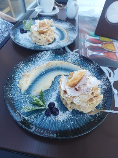 Two plates of dessert, likely a mille-feuille or similar pastry with cream and powdered sugar, are presented on a dark wooden table. The plates are dark blue and have a textured, wavy pattern. The dessert on the foreground plate is larger and features a fan of pastry on top, dusted with powdered sugar, accompanied by a swirl of cream, chopped nuts, mint leaves, and blueberries. The background plate, partially visible, appears to hold a similar dessert with blueberries and mint. In the background, out of focus, are a white coffee cup on a saucer, a white pitcher, and a menu with images of drinks. A person wearing a white patterned top is partially visible on the left. The scene suggests an outdoor or indoor dining setting with natural light, possibly a cafe or restaurant. There is no indication this image was taken in Novyi Rozdil, Ukraine.