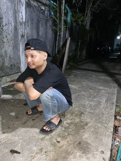 A young man wearing a black t-shirt, blue jeans, a black baseball cap, and black slides is crouching on a concrete walkway. The walkway is gritty and has some debris on it, including leaves and what appears to be a small piece of wood. To the left of the man is a rough, weathered concrete wall. To his right, the walkway extends into a darker area with trees and foliage. In the background, a car with its headlights on is visible on a darker path, suggesting this is an outdoor urban setting. The lighting suggests it is nighttime or early evening. The overall scene conveys a casual, possibly outdoor waiting or resting scenario. The location appears to be a residential or less-trafficked area within Banjarmasin, Indonesia, given the tropical vegetation and the style of the wall and walkway.