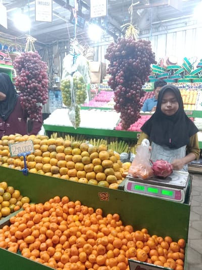 This image depicts the interior of a brightly lit fruit market stall located in Sekopek Wetan, Indonesia. The scene is dominated by tiered green display shelves laden with hundreds of small, spherical orange citrus fruits. A yellow price tag displaying "28.000" is attached to the upper tier of these fruits. Three large clusters of grapes hang from the ceiling: two are dark red/purple, and one in the center consists of green grapes contained within clear plastic bags.
In the foreground to the right, a young woman, likely a vendor, is visible from the waist up. She wears a black hijab, a light-colored long-sleeved top, and blue denim overalls. Her gaze is directed downwards towards a white and pink digital weighing scale. On the scale platform, a clear plastic bag filled with orange fruits (similar to those on display) rests next to a single red dragon fruit. The scale's digital displays show green numbers.
In the mid-ground to the left, a second woman, also wearing a black hijab and a dark maroon garment, is partially visible with her back mostly to the viewer. In the background, behind the main fruit displays, a man wearing a light blue polo shirt is partially visible. Additional fruit displays are present in the background, including rows of yellow bananas, heaps of red, spiky fruits (likely rambutan), and other lighter-colored rounded fruits. Overhead, fluorescent lights illuminate the stall, and white signs, one partially reading "INOVASI," are suspended from the ceiling structure.