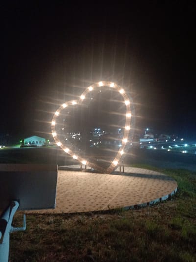 An illuminated heart-shaped sculpture is featured in the foreground, with its outline comprised of numerous small lights. The sculpture sits atop a circular, patterned paved area. To the left of the paved area, a portion of a grey, outdoor seating structure is visible. Behind the sculpture and the paved area, a grassy expanse is present. In the middle ground, a white tent-like structure with arched openings and green lighting is situated. Beyond the tent, a blurred cityscape with scattered lights, streetlights, and buildings can be observed, suggesting an urban or semi-urban environment. The overall scene is captured at night, with the dark sky and the illuminated heart sculpture being the prominent elements. This image was taken in Zarubino, Russia.