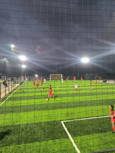 A fenced soccer field with artificial turf is lit by overhead lights. Several young players in red and white uniforms are practicing on the field. A few individuals are on the sidelines, and a parked vehicle is visible in the distance. The view is partially obstructed by the fence netting. The sky is dark, and there appears to be a cloud formation resembling a face.