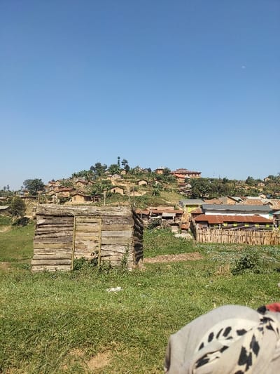 The image depicts a rural, hillside settlement in Kitsombiro, Democratic Republic of the Congo, under a clear blue sky with a faint crescent moon visible in the upper right. In the immediate foreground, the top of a person's shoulder and head are partially visible, clad in a light grey garment with a black and white patterned fabric. The ground in the foreground consists primarily of green grass with patches of exposed earth.
Central to the midground is a rectangular, single-story structure constructed from horizontally stacked raw logs or rough-hewn timber, appearing to be a shed or small building. Its front face includes vertical planks forming a door or frame. To the right of this wooden structure, the landscape transitions into a cluster of additional buildings and cultivated areas. Notable are several low-rise structures with corrugated metal roofs, some reddish-brown, some grey, often surrounded by wooden picket fences. One longer building features a reddish-brown corrugated roof and distinct green-painted elements along its walls, also enclosed by a wooden fence.
The background is dominated by a steeply rising hill densely covered with numerous small, simple dwellings. These structures exhibit a variety of building materials, including earthen walls, thatched roofs, and corrugated metal roofs of various colors. Trees and other vegetation are interspersed throughout the settlement on the hillside. At the highest point of the hill, a larger, multi-story building with a reddish-brown roof and visible windows stands out. No discernible human activity or other specific subjects are evident beyond the architectural elements and natural environment.