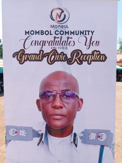 The image is of a white banner with black lettering that reads, “Mombol Community Congratulates You on Your Grand Civic Reception.” The banner features a large photo of a man in a uniform with a badge. The man is wearing glasses and has a serious expression. The banner is likely located in Mombol, Nigeria, at the coordinates 10.116 latitude, 13.046 longitude. The background appears to be a dirt lot.