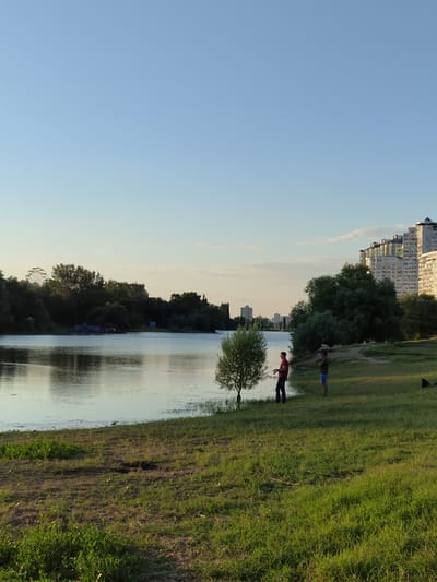 The image depicts a serene riverside scene in Novomoskovsk, Russia, likely during the late afternoon or early evening, given the soft, warm light. The foreground is dominated by a grassy embankment sloping down towards a calm body of water, possibly a river or lake. Lush green grass covers the ground, with patches of dry earth visible.
On the right side of the embankment, two individuals are present. One person, wearing a red shirt and dark pants, appears to be fishing, holding a fishing rod. Standing near them, to their right, is another person dressed in a blue shirt and shorts, also facing the water. A small, solitary tree stands on the edge of the water between them.
In the background, across the water, a line of trees forms a dark green border, with a Ferris wheel faintly visible in the distance to the left. Further in the background, a cluster of modern, multi-story apartment buildings rises against a clear, pale blue sky. The water reflects the sky and the surrounding greenery, creating a peaceful, tranquil atmosphere. The overall impression is one of a quiet, suburban recreational area.