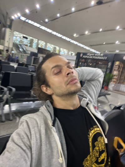 The photograph appears to have been taken in an airport terminal. A man with light skin, brown hair, and stubble lies sideways on chairs. He is wearing a grey hoodie over a black t-shirt with a gold design. The terminal has rows of unoccupied seating, and in the background, there's a sign that says "Popular Pharmacy". The location is likely Lal Bahadur Shastri International Airport, Babatpur, India.