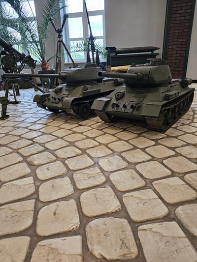 The image shows a T-80 tank and other military equipment on display at the Vadim Zadorozhny Technology Museum in Arkhangel’skoye, Russia. The tank is olive green and positioned on a textured surface resembling cobblestone. Behind the tank, there are windows with open sections, foliage from what appears to be a palm tree, and other military artifacts. A brick wall is visible in the background.