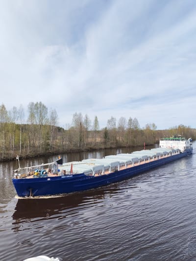 A long blue freighter, "Павел Лебедев" (Pavel Lebedev), is navigating a waterway. The freighter is loaded with containers and has a white superstructure at the stern. The vessel is moving on a body of water, most likely a canal or river, and the surrounding environment consists of trees on the banks. The location is Srednii Rubezh, Russia. The sky is partly cloudy.