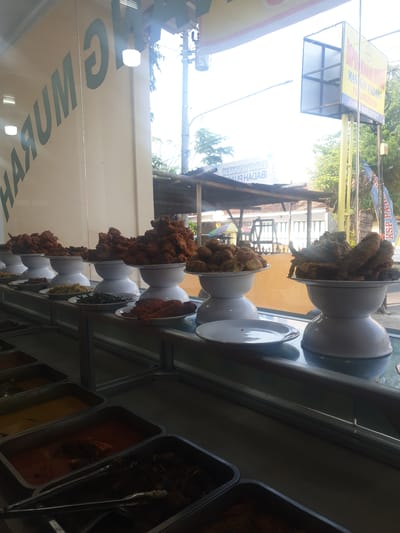 The image shows a display of Indonesian food, likely at a roadside eatery or a small restaurant. The food is arranged in white bowls and metal trays, with many of the dishes appearing to be fried. These include what looks like fried chicken pieces and perhaps some fried vegetables.

The setting appears to be an indoor space with a large window or glass front, through which the outside is visible. The exterior shows trees, buildings, and what looks like a street. There's a bright, sunny day suggested by the strong light coming through the window, causing some glare.

Text is visible on a yellow sign in the upper left portion of the image. It appears to be a business sign. Another sign with Indonesian text is reflected on the glass in the lower left. The reflection reads "MASAKAN JAMU HASMADA".

The overall impression is of a bustling food stall or a casual eatery ready to serve customers, with a variety of popular Indonesian dishes on display. The focus is on the food itself, presented in an appetizing manner.