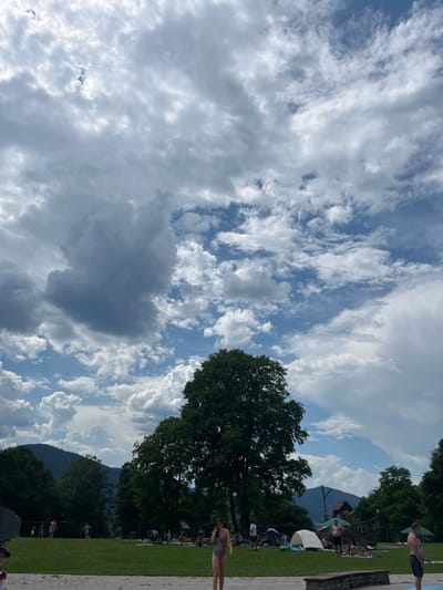 The image depicts an outdoor scene at a swimming pool complex in Oberammergau, Germany. The sky is filled with dramatic clouds, with patches of blue visible. Several people are scattered across a grassy area, some sunbathing on towels, others standing or walking. In the foreground, a paved area suggests the edge of a pool. Beyond the grassy area, there are trees and in the background, mountains are visible. Some recreational structures, possibly a playground, are also present. The overall impression is a relaxed, summery atmosphere.