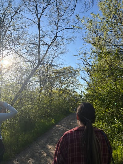 The image depicts a low-angle view of a person on a dirt path in Staraya Stanitsa, Russia. The sky is visible through bare tree branches on the left and foliage overhead. The person wears a red plaid shirt and their hair is pulled back. A path runs alongside a grassy area with sunlight filtering through.
