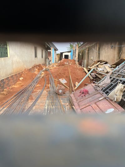 In Edopa, Nigeria, a construction site is depicted with a focus on building materials. A pile of red soil sits prominently, alongside bundles of metal rebar. Various construction debris, including wooden planks and a metal frame, are scattered around the area. A large wooden door lies on the ground, and a beige backpack is placed on top of the metal frame. The scene appears to be situated between two buildings, with one on the left featuring a light-colored stucco exterior and a green-barred window, and the other on the right showing a concrete wall.
